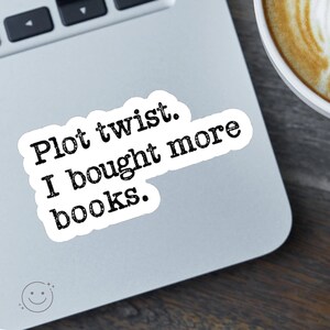 May include: A white sticker with the text "Plot twist. I bought more books." in black, placed on a laptop. A cup of coffee is visible in the background. The sticker has a rounded rectangle shape.