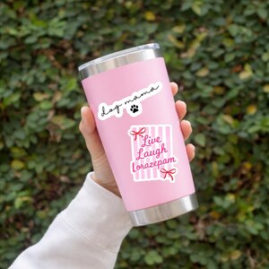 May include: A pink stainless steel tumbler with a silver rim and base. The tumbler has two stickers: one that says "dog mama" with a paw print, and another that says "Live Laugh Lorazepam" with red bows and pink stripes.