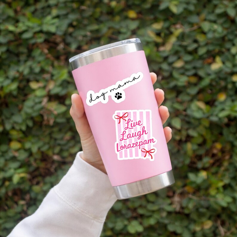 May include: A pink stainless steel tumbler with a silver rim and base. The tumbler has two stickers: one that says "dog mama" with a paw print, and another that says "Live Laugh Lorazepam" with red bows and pink stripes.