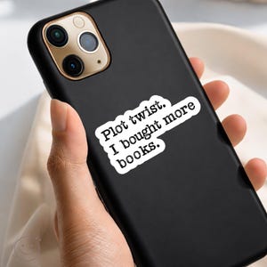 May include: Black phone case with a white sticker that reads "Plot twist. I bought more books." The phone case has a gold camera ring and is held in a person's hand. The background is a light beige color.