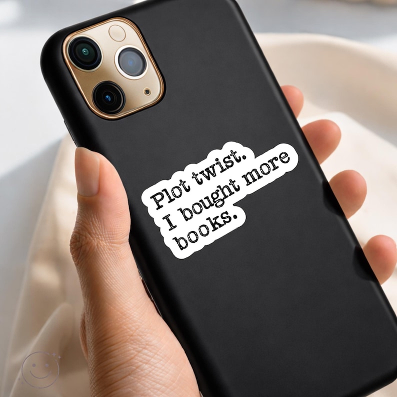 May include: Black phone case with a white sticker that reads "Plot twist. I bought more books." The phone case has a gold camera ring and is held in a person's hand. The background is a light beige color.