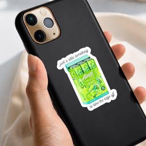 K&ouml;nnte beinhalten: Ein schwarzes Smartphone mit einem Aufkleber eines limettengr&uuml;nen Alani Nu Energy-Drink-Packs. Der Aufkleber enth&auml;lt den Text "Just a little something" &uuml;ber den Dosen und "to take the edge off" darunter, mit dem Aufdruck "Lime Slush" auf den Dosen.