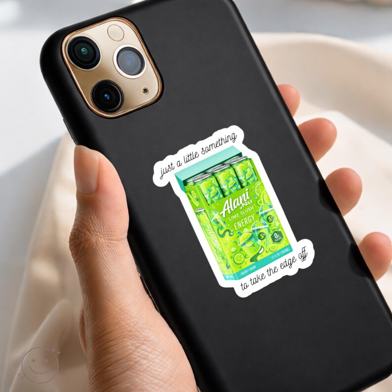 K&ouml;nnte beinhalten: Ein schwarzes Smartphone mit einem Aufkleber eines limettengr&uuml;nen Alani Nu Energy-Drink-Packs. Der Aufkleber enth&auml;lt den Text "Just a little something" &uuml;ber den Dosen und "to take the edge off" darunter, mit dem Aufdruck "Lime Slush" auf den Dosen.