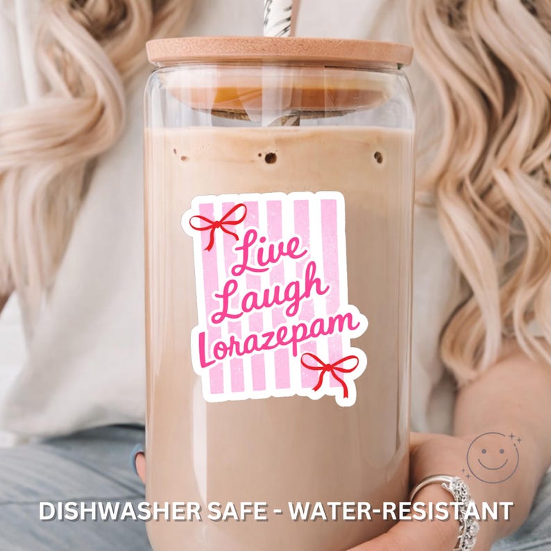 May include: A clear glass beverage cup with a wooden lid and a straw. A pink and white sticker on the cup reads "Live Laugh Lorazepam" in cursive script. The sticker has red bow accents. The cup is filled with a light brown liquid.