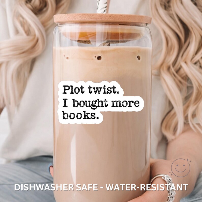 May include: A clear glass tumbler with a wooden lid and a straw, filled with a light brown beverage. The tumbler has a white sticker with the text "Plot twist. I bought more books." The words "Dishwasher Safe - Water-Resistant" are at the bottom.