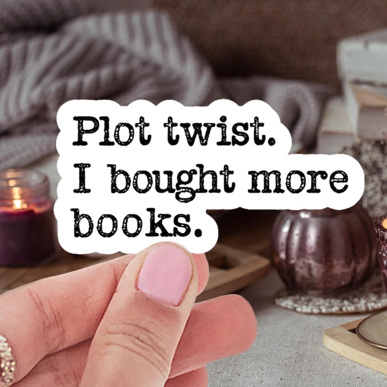 May include: A white sticker with black text that reads "Plot twist. I bought more books." is held by a person. The background includes a candle, a decorative pumpkin, and a stack of books, suggesting a cozy reading theme.
