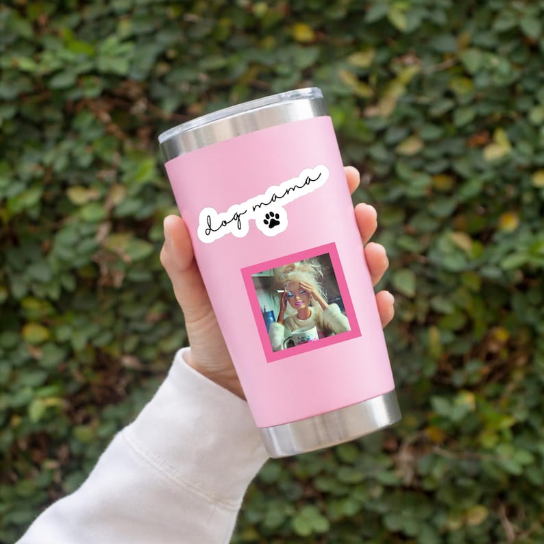 May include: A light pink stainless steel tumbler with a silver rim and base. The tumbler features a white sticker with the black text "dog mama" and a small paw print icon, along with a square photograph of a blonde doll holding a small cup.