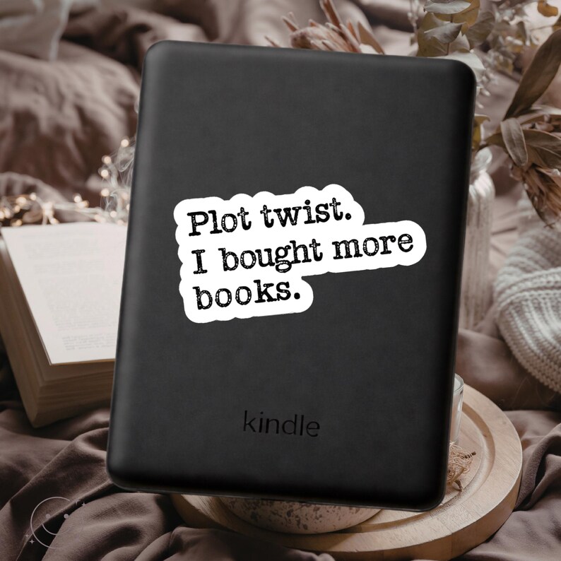 May include: A black Kindle e-reader with a white sticker that reads "Plot twist. I bought more books." The word "kindle" is printed at the bottom. The e-reader is on a brown surface.
