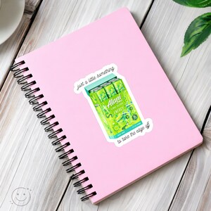 K&ouml;nnte beinhalten: Ein hellrosa Notizbuch mit Spiralbindung und einem Aufkleber eines limettengr&uuml;nen Alani Nu Energy-Drink-Packs. Der Aufkleber enth&auml;lt den Text "just a little something" &uuml;ber den Dosen und "to take the edge off" unter den Dosen in schwarzer Schreibschrift.