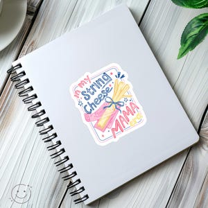 May include: A sticker with a watercolour illustration of string cheese, with the text "In my String Cheese Mama Era" in pink and blue. The sticker is on a white spiral-bound notebook.