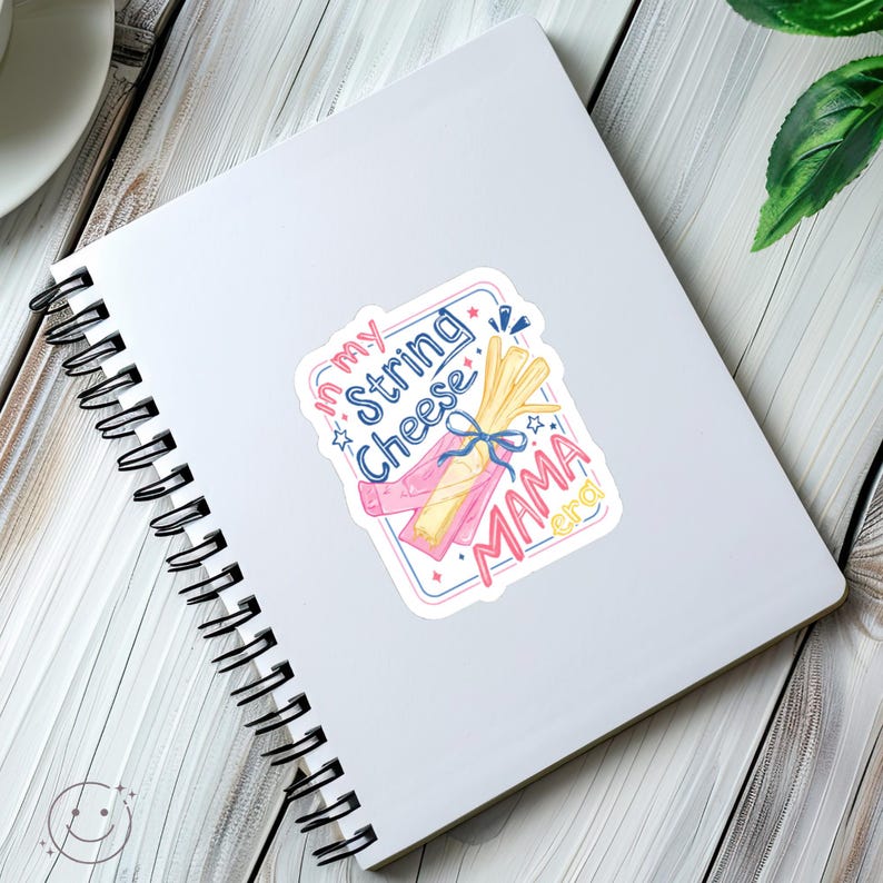 May include: A sticker with a watercolour illustration of string cheese, with the text "In my String Cheese Mama Era" in pink and blue. The sticker is on a white spiral-bound notebook.