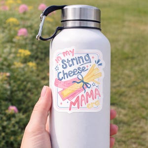 May include: A white water bottle with a silver lid and a black handle. A sticker on the bottle reads "In my String Cheese Mama era" with illustrations of string cheese and a bow. The background is a blurred green and pink.