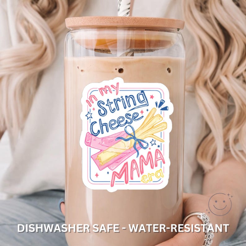 May include: A clear glass tumbler with a wooden lid and a straw, filled with a creamy beverage. The tumbler has a sticker with the text "In my String Cheese Mama Era" and a string cheese graphic. The words "Dishwasher Safe - Water-Resistant" are also visible.