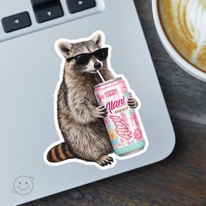 May include: A die-cut sticker featuring a brown and grey raccoon wearing black sunglasses and drinking from a pink can labeled Cotton Candy Alani Energy with a straw. The raccoon has a striped tail and is shown in a standing position.