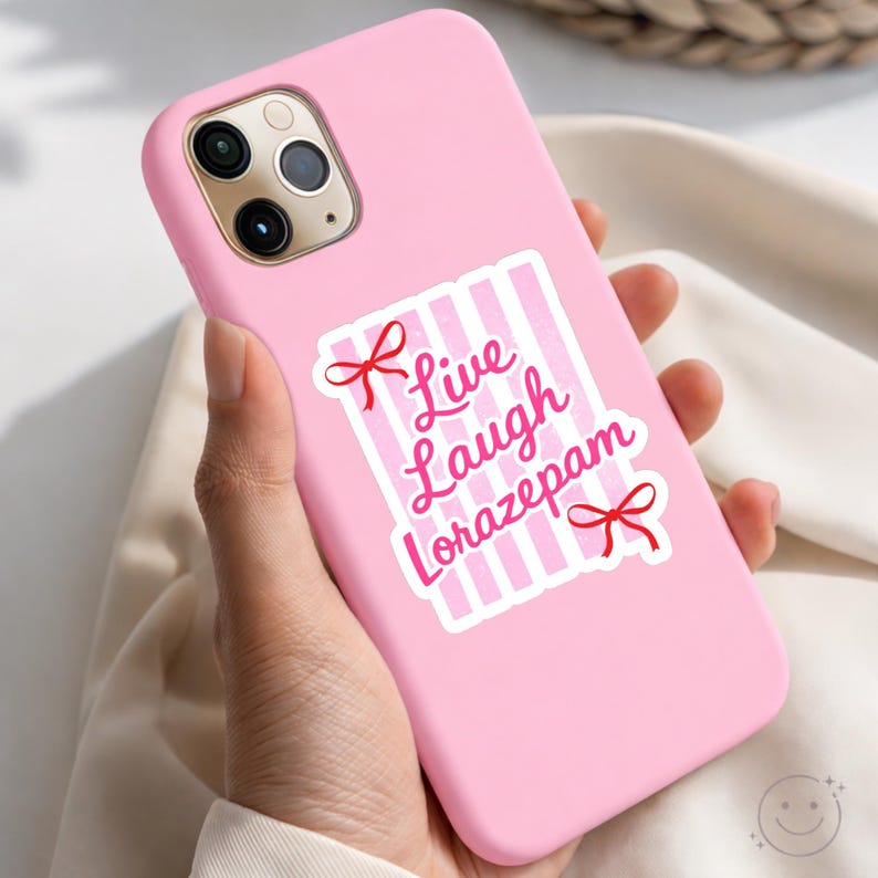 May include: Pink phone case with a sticker that reads "Live Laugh Lorazepam" in pink script. The sticker has a white background with pink vertical stripes and red bow accents. The phone case has a smooth, matte finish.