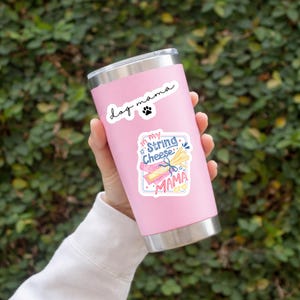 May include: A pink stainless steel travel mug with a silver rim and lid. The mug has two stickers: one that says "Dog mama" with a paw print, and another that says "my String Cheese mama" with a cheese illustration.