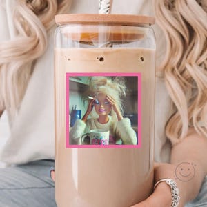 May include: A clear glass tumbler with a bamboo lid and a straw, filled with a light brown iced coffee. A square sticker on the front features a blonde doll with messy hair, holding a cigarette, and resting her hands on her head near a mug.