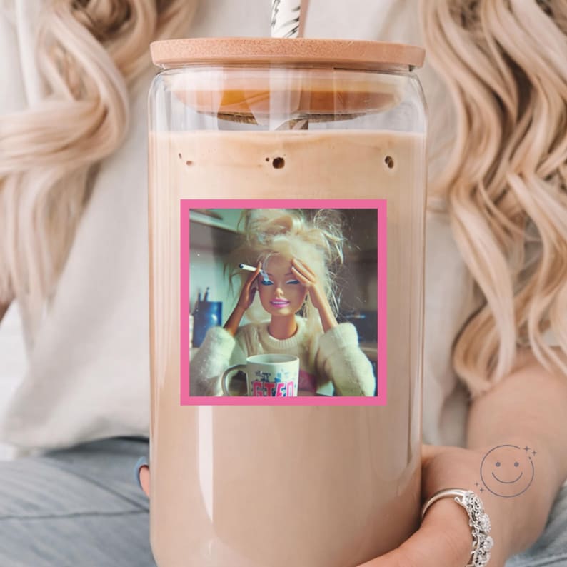 May include: A clear glass tumbler with a bamboo lid and a straw, filled with a light brown iced coffee. A square sticker on the front features a blonde doll with messy hair, holding a cigarette, and resting her hands on her head near a mug.
