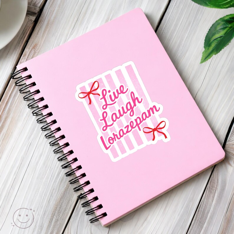 May include: A pink spiral-bound notebook with a sticker that reads "Live Laugh Lorazepam" in pink script. The sticker has a striped background and red bow accents. The notebook is on a white wooden surface.