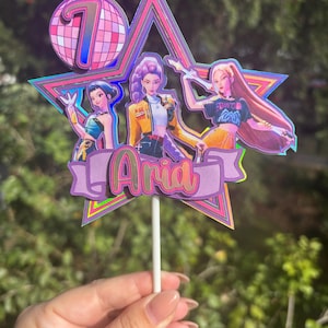 May include: A cake topper with three cartoon figures, a disco ball with the number 7, and the name "Aria". The topper is multi-layered with a star shape and a purple banner. The figures are in various poses.