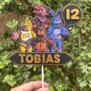 Könnte beinhalten: Ein farbenfroher Tortenaufsatz mit Figuren aus einem beliebten Videospiel. Der Aufsatz zeigt den Namen "TOBIAS" und die Zahl "12". Die Figuren sind in Gelb, Rot, Braun und Blau gehalten. Der Aufsatz wird von einem weißen Stab gehalten.