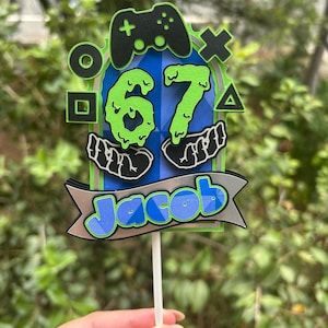 Puede incluir: Adorno para tarta con temática de videojuegos. Presenta un mando negro, el número 67 en verde y el nombre "Jacob" en azul. El adorno está sobre un palito blanco y tiene una combinación de colores azul y verde.