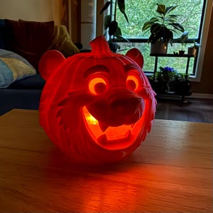 Può includere: Un secchiello a forma di zucca in plastica arancione brillante e illuminato, con le sembianze di un muso di tigre con orecchie arrotondate, occhi spalancati e un sorriso dentato. La luce interna illumina i dettagli intagliati.
