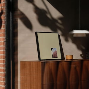 May include: A framed picture with a black border, featuring a small image of a building, rests on a wooden dresser. A small, round wooden container sits next to the frame. The scene is lit by natural light.