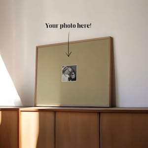 May include: A framed photograph with a light wood frame and a beige mount. The photograph is a small, black and white image. The text "Your photo here!" is above the photograph. The frame is resting on a wooden cabinet.