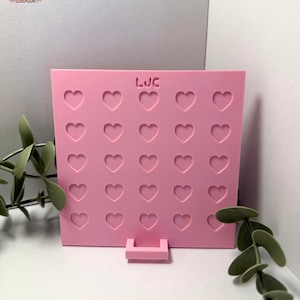 May include: A pink, heart-shaped display board with 25 cut-out hearts. The board has a small rectangular base and the letters "LJC" are visible at the top. The board is made of a solid material and is suitable for displaying small items.