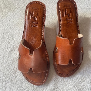Peut inclure: Une paire de sandales plates en cuir marron avec une large bride en forme de H sur le dessus. La semelle int&eacute;rieure est estampill&eacute;e du texte VRAI CUIR et MAROC avec un symbole de main. Les sandales pr&eacute;sentent des coutures visibles sur le bord.