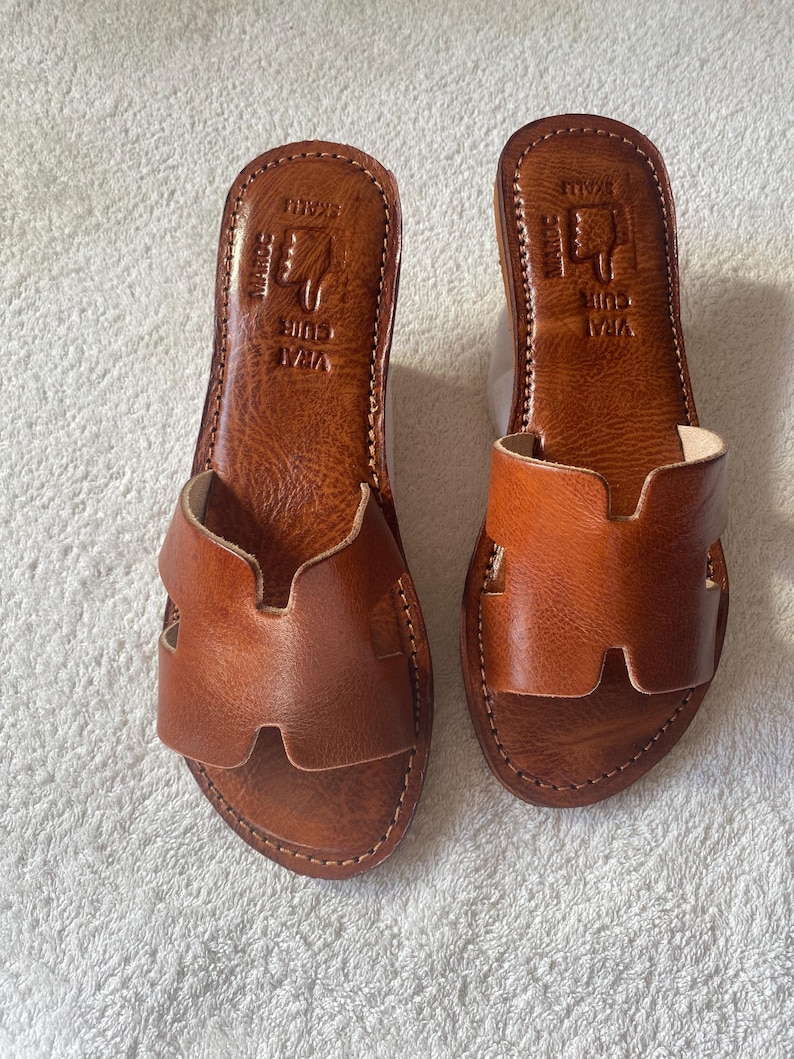 Peut inclure: Une paire de sandales plates en cuir marron avec une large bride en forme de H sur le dessus. La semelle int&eacute;rieure est estampill&eacute;e du texte VRAI CUIR et MAROC avec un symbole de main. Les sandales pr&eacute;sentent des coutures visibles sur le bord.
