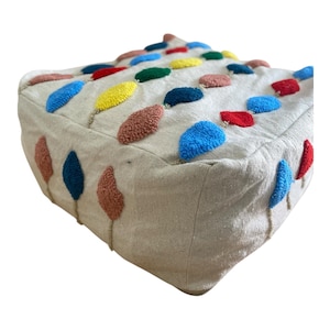 Op de afbeelding: Een vierkant vloerkussen van gebroken wit textiel met een getuft, gestructureerd patroon van kleine, ruitvormige plukjes in levendige kleuren zoals rood, blauw, geel, groen en terracotta, gerangschikt in een raster over de bovenkant en de zijkanten.