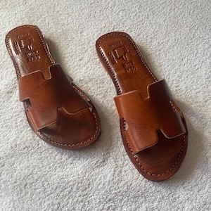 Peut inclure: Une paire de sandales plates en cuir marron avec une bride large en forme de H sur le dessus. La semelle int&eacute;rieure porte une estampe avec le texte VRAI CUIR et une ic&ocirc;ne de main. Les bords des semelles pr&eacute;sentent des coutures apparentes.