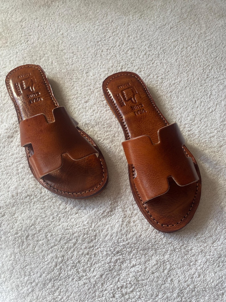 Peut inclure: Une paire de sandales plates en cuir marron avec une bride large en forme de H sur le dessus. La semelle int&eacute;rieure porte une estampe avec le texte VRAI CUIR et une ic&ocirc;ne de main. Les bords des semelles pr&eacute;sentent des coutures apparentes.