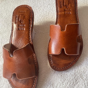 Peut inclure: Une paire de sandales plates en cuir marron clair avec une large bride en forme de H sur le dessus. La semelle int&eacute;rieure est estampill&eacute;e du texte VRAI CUIR et MAROC avec une ic&ocirc;ne de main. Les bords pr&eacute;sentent des coutures apparentes.