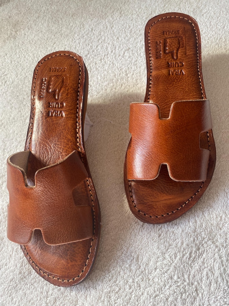 Peut inclure: Une paire de sandales plates en cuir marron clair avec une large bride en forme de H sur le dessus. La semelle int&eacute;rieure est estampill&eacute;e du texte VRAI CUIR et MAROC avec une ic&ocirc;ne de main. Les bords pr&eacute;sentent des coutures apparentes.