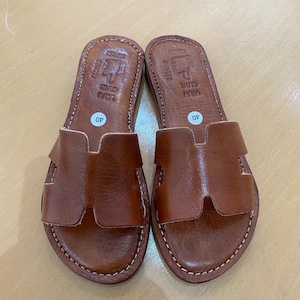 "A pair of artisan handcrafted Moroccan leather flat sandals featuring a classic -style strap. Made from high-quality natural grain leather with meticulous hand-stitching, shown from a top-down view on a neutral surface."
