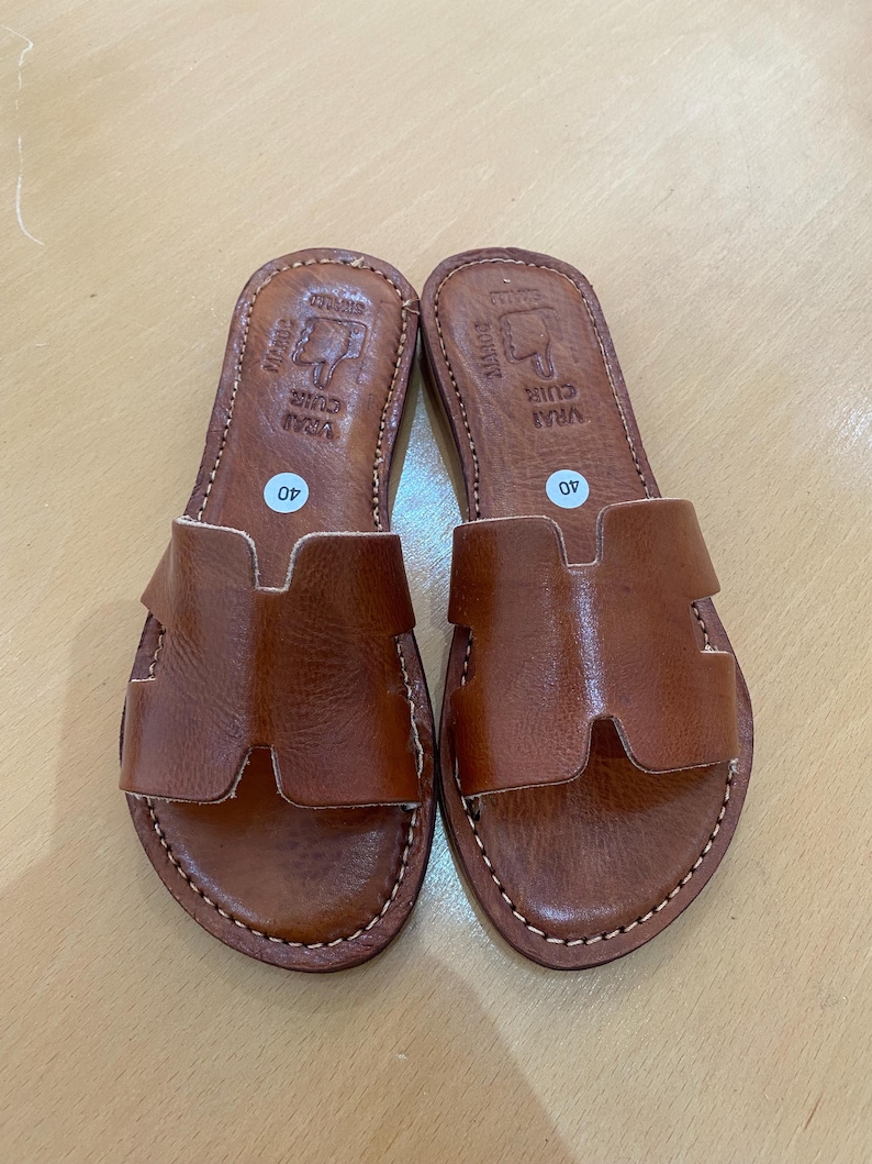 "A pair of artisan handcrafted Moroccan leather flat sandals featuring a classic -style strap. Made from high-quality natural grain leather with meticulous hand-stitching, shown from a top-down view on a neutral surface."