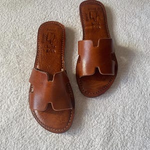 Peut inclure: Une paire de sandales plates en cuir marron avec une large bride en forme de H sur le dessus. La semelle int&eacute;rieure porte l'inscription gaufr&eacute;e VRAI CUIR et SKALLE. Les sandales pr&eacute;sentent des coutures apparentes sur tout le contour.