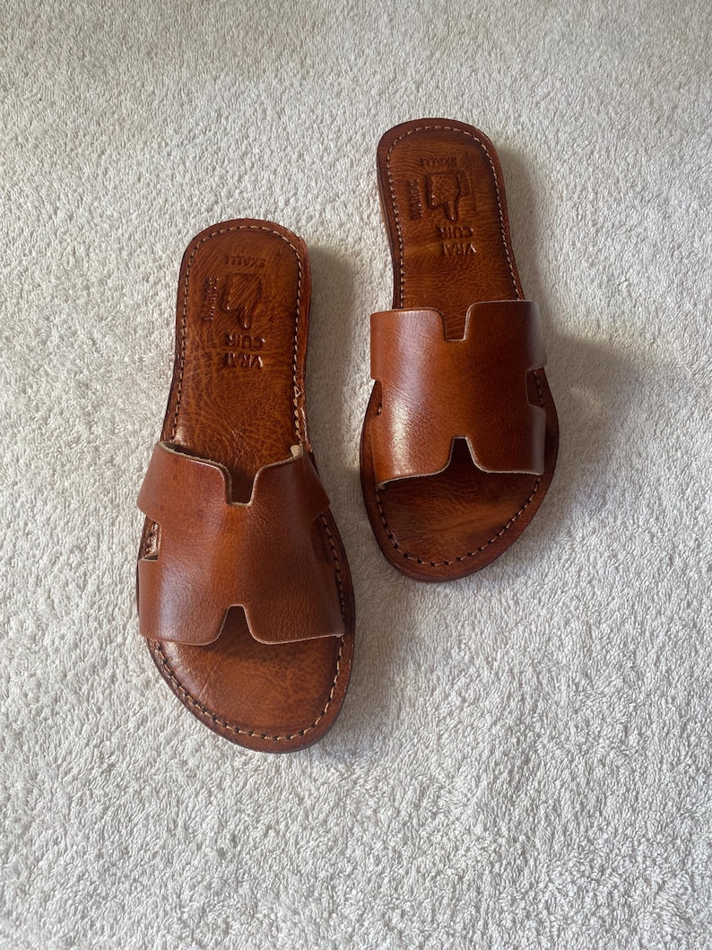 Peut inclure: Une paire de sandales plates en cuir marron avec une large bride en forme de H sur le dessus. La semelle int&eacute;rieure porte l'inscription gaufr&eacute;e VRAI CUIR et SKALLE. Les sandales pr&eacute;sentent des coutures apparentes sur tout le contour.
