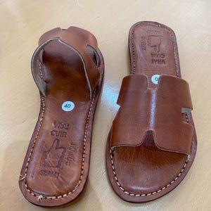 "A pair of artisan handcrafted Moroccan leather flat sandals featuring a classic -style strap. Made from high-quality natural grain leather with meticulous hand-stitching, shown from a top-down view on a neutral surface."