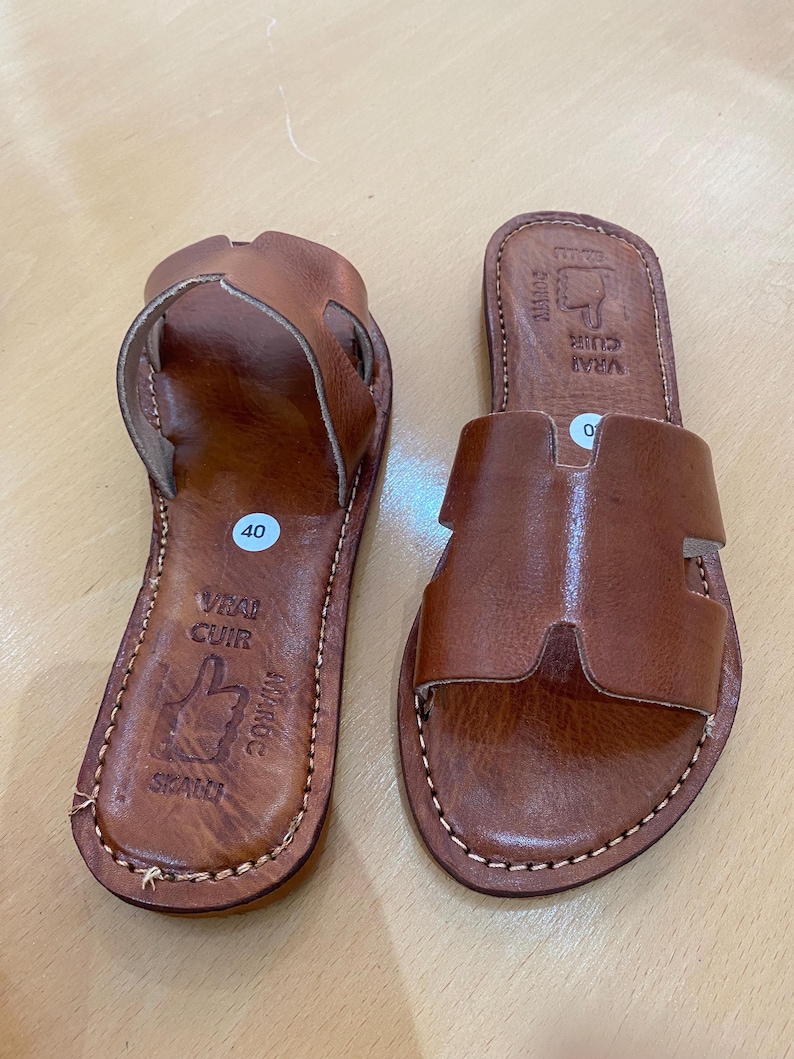 "A pair of artisan handcrafted Moroccan leather flat sandals featuring a classic -style strap. Made from high-quality natural grain leather with meticulous hand-stitching, shown from a top-down view on a neutral surface."