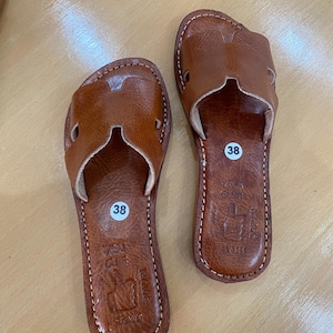 "A pair of artisan handcrafted Moroccan leather flat sandals featuring a classic -style strap. Made from high-quality natural grain leather with meticulous hand-stitching, shown from a top-down view on a neutral surface."