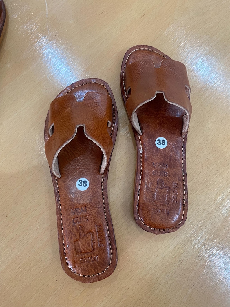 "A pair of artisan handcrafted Moroccan leather flat sandals featuring a classic -style strap. Made from high-quality natural grain leather with meticulous hand-stitching, shown from a top-down view on a neutral surface."