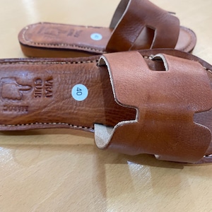"A pair of artisan handcrafted Moroccan leather flat sandals featuring a classic -style strap. Made from high-quality natural grain leather with meticulous hand-stitching, shown from a top-down view on a neutral surface."