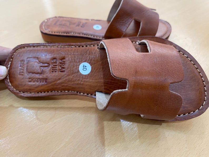 "A pair of artisan handcrafted Moroccan leather flat sandals featuring a classic -style strap. Made from high-quality natural grain leather with meticulous hand-stitching, shown from a top-down view on a neutral surface."