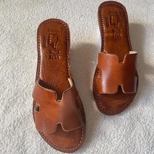 Peut inclure: Une paire de sandales plates en cuir marron avec une large bride en forme de H sur le dessus. La semelle int&eacute;rieure est estampill&eacute;e avec le texte VRAI CUIR, MAROC et SKALLI, ainsi qu'une ic&ocirc;ne de main. Les sandales pr&eacute;sentent des coutures visibles.