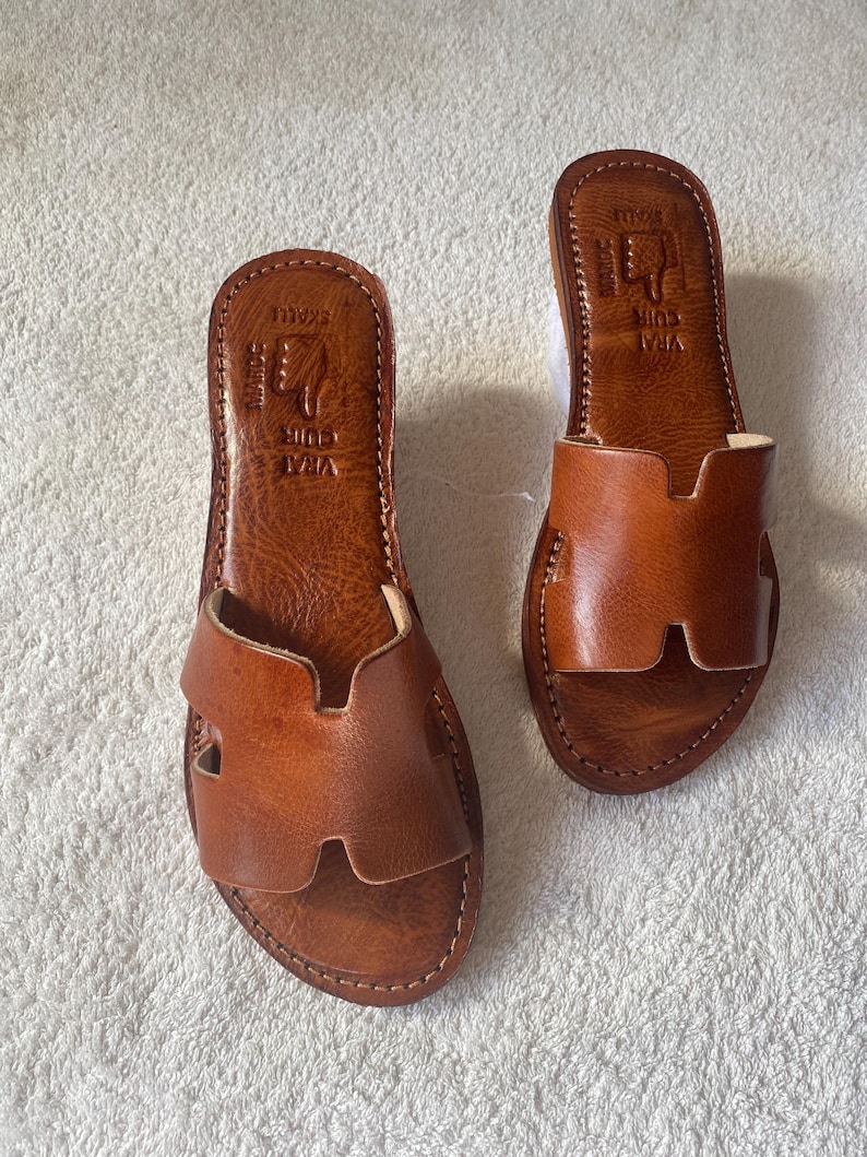 Peut inclure: Une paire de sandales plates en cuir marron avec une large bride en forme de H sur le dessus. La semelle int&eacute;rieure est estampill&eacute;e avec le texte VRAI CUIR, MAROC et SKALLI, ainsi qu'une ic&ocirc;ne de main. Les sandales pr&eacute;sentent des coutures visibles.