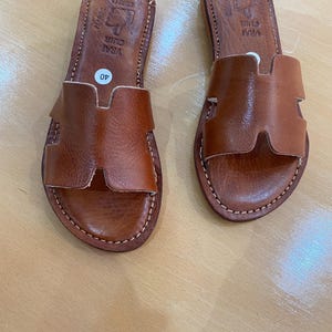 "A pair of artisan handcrafted Moroccan leather flat sandals featuring a classic -style strap. Made from high-quality natural grain leather with meticulous hand-stitching, shown from a top-down view on a neutral surface."